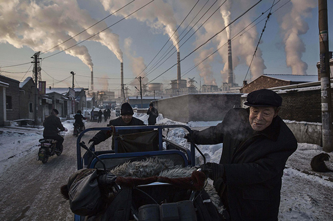 China's Coal Addiction - Kevin Frayer - Smoke billows from stacks as men push a tricycle through a neighborhood next to a coal-fired power plant in northern Shanxi province.